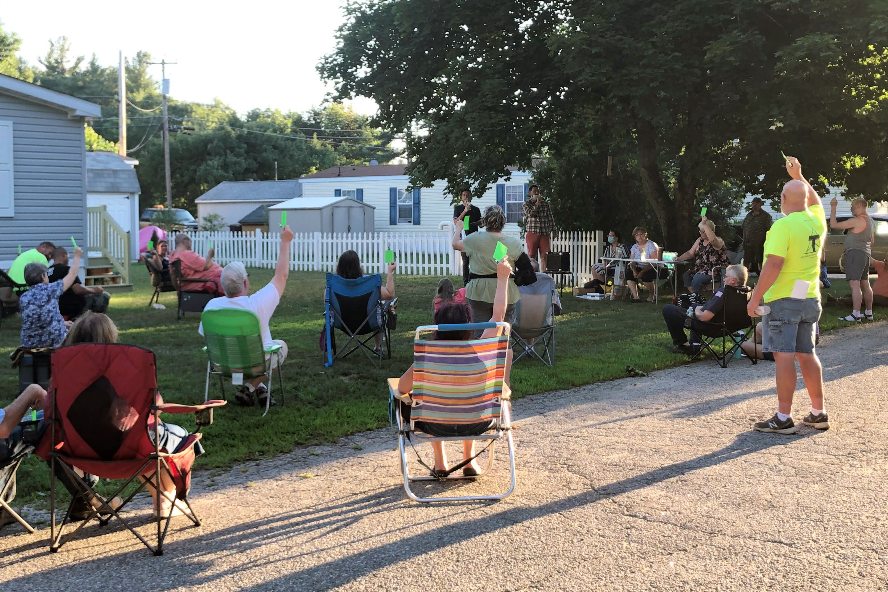 A group of people holding their arms up to vote on a lawn in a manufactured home park.