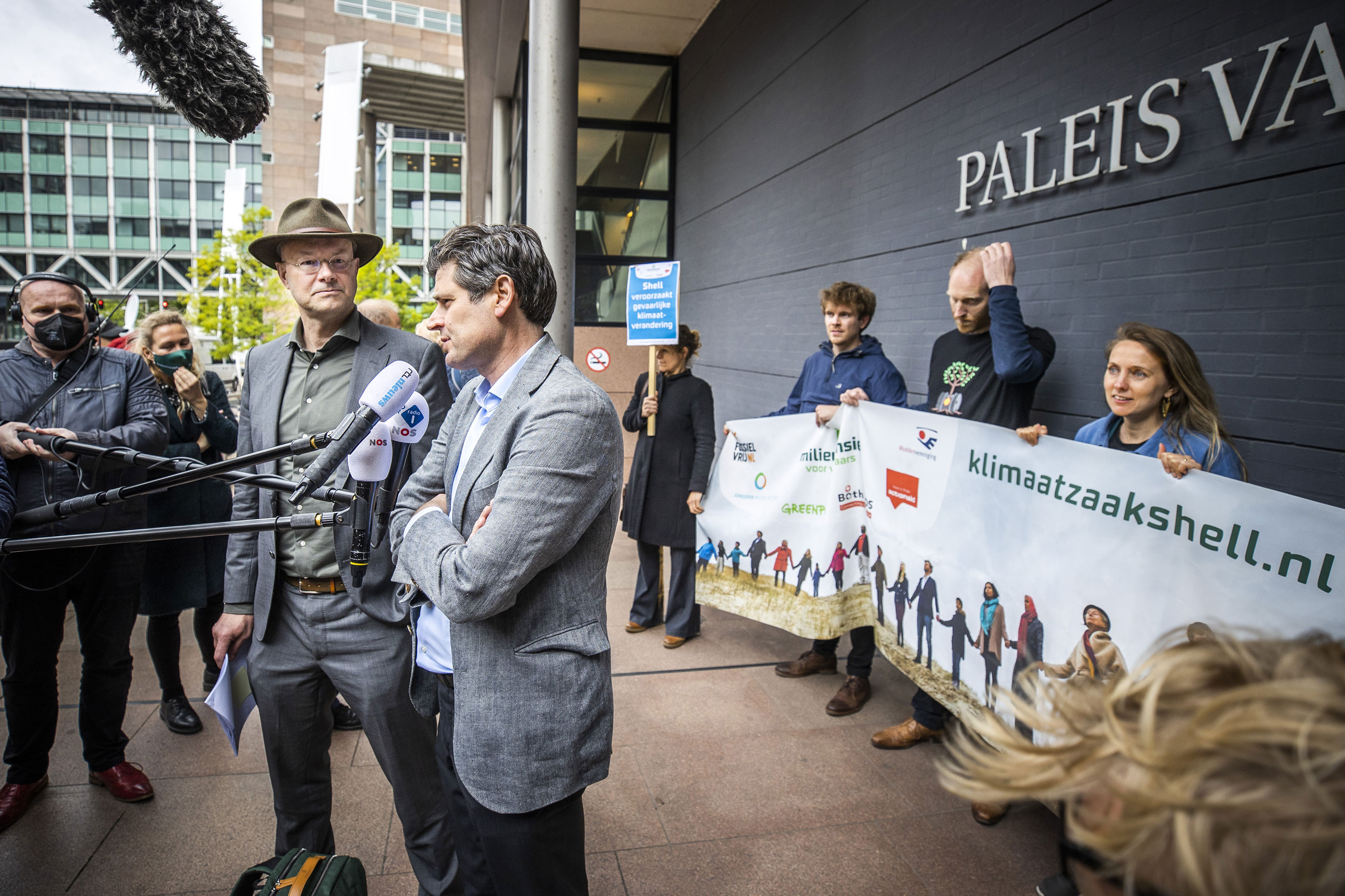 Two people speaking in front of microphones in front of a group of people holding a banner